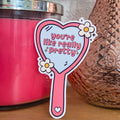 A die-cut sticker with a cute mirror design that reads 'You're Like Really Pretty’ with daisies on the sides resting on a wooden surface.