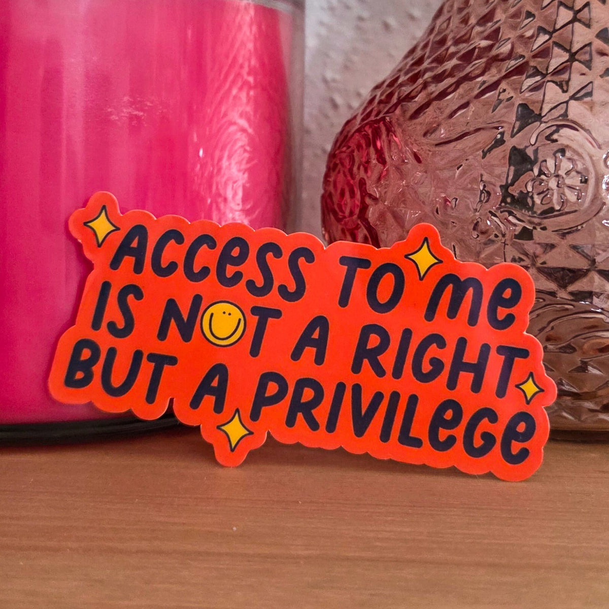 A bright die-cut sticker with an orange design with a smiley face and stars that reads 'access to me is not a right but a privilege’ resting on a wooden surface.