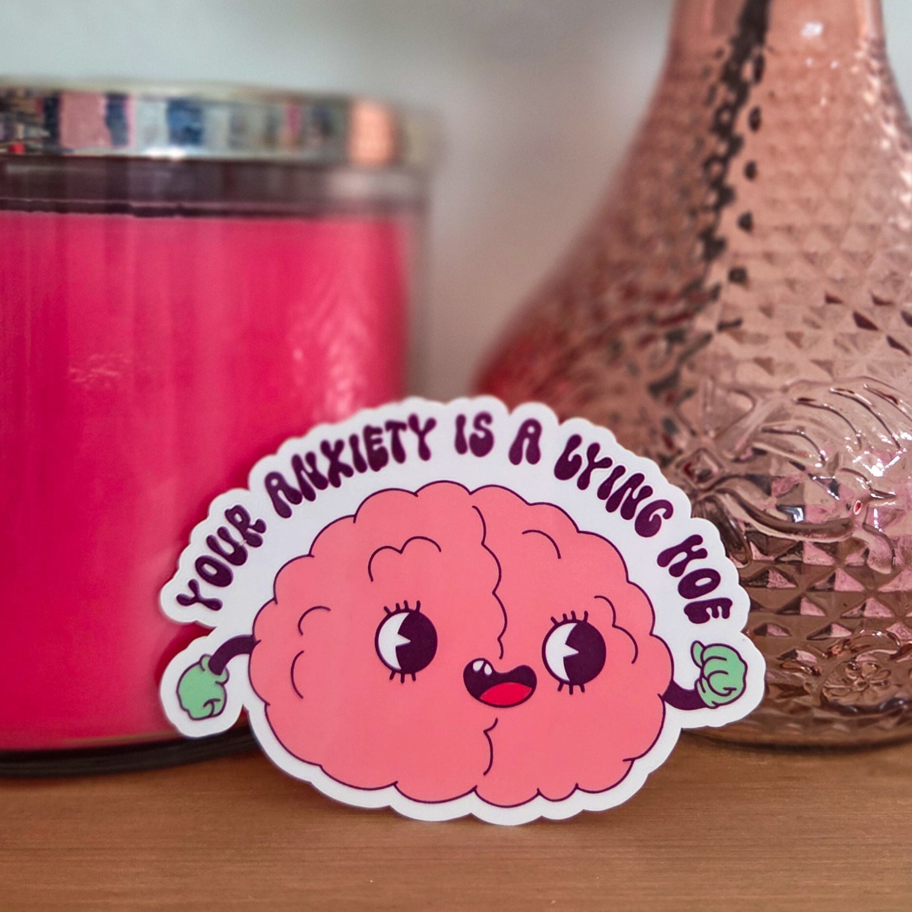 Pink brain-shaped sticker with text next to a pink candle and vase on a wooden surface.