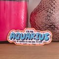 A novelty sticker with a wavy border and the text 'I'M AN AQUARIUS OF COURSE I'M TOO COOL TO CARE' in blue, pink, and white, displayed on a wooden surface with a pink candle and a textured vase in the background.