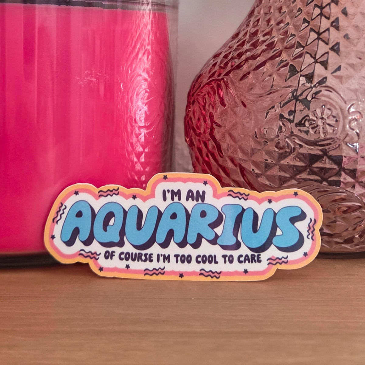 A novelty sticker with a wavy border and the text 'I'M AN AQUARIUS OF COURSE I'M TOO COOL TO CARE' in blue, pink, and white, displayed on a wooden surface with a pink candle and a textured vase in the background.