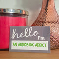 Pink candle, vase with peach rose, and 'hello I'm an audiobook addict' sign on a wooden surface.