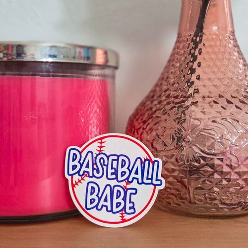 Pink candle and textured vase with 'Baseball Babe' sticker on a wooden surface