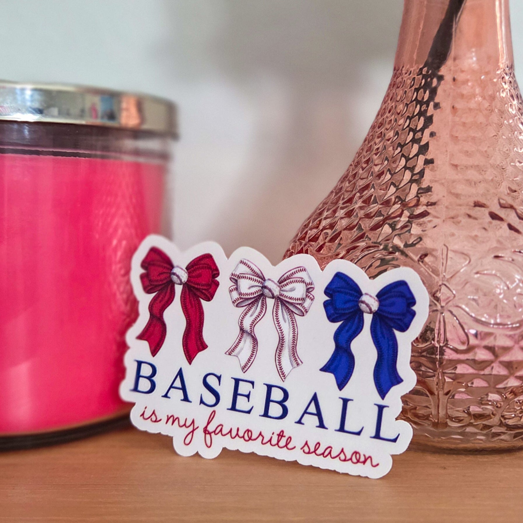 Sticker with 'Baseball is my favorite season' text on a wooden surface with a pink candle and vase in the background.