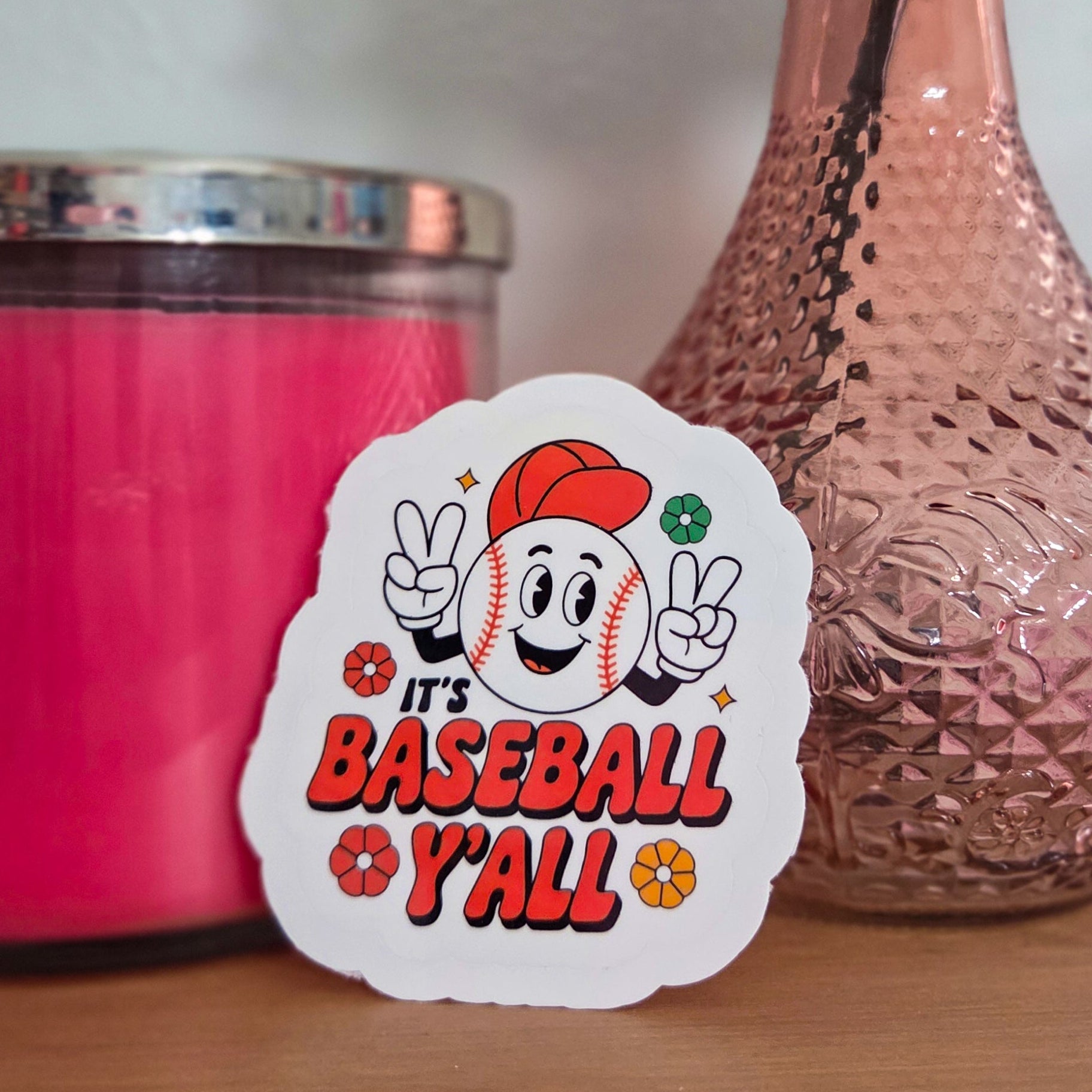 Sticker with 'It's Baseball Y'all' design next to a pink candle and vase on a wooden surface.