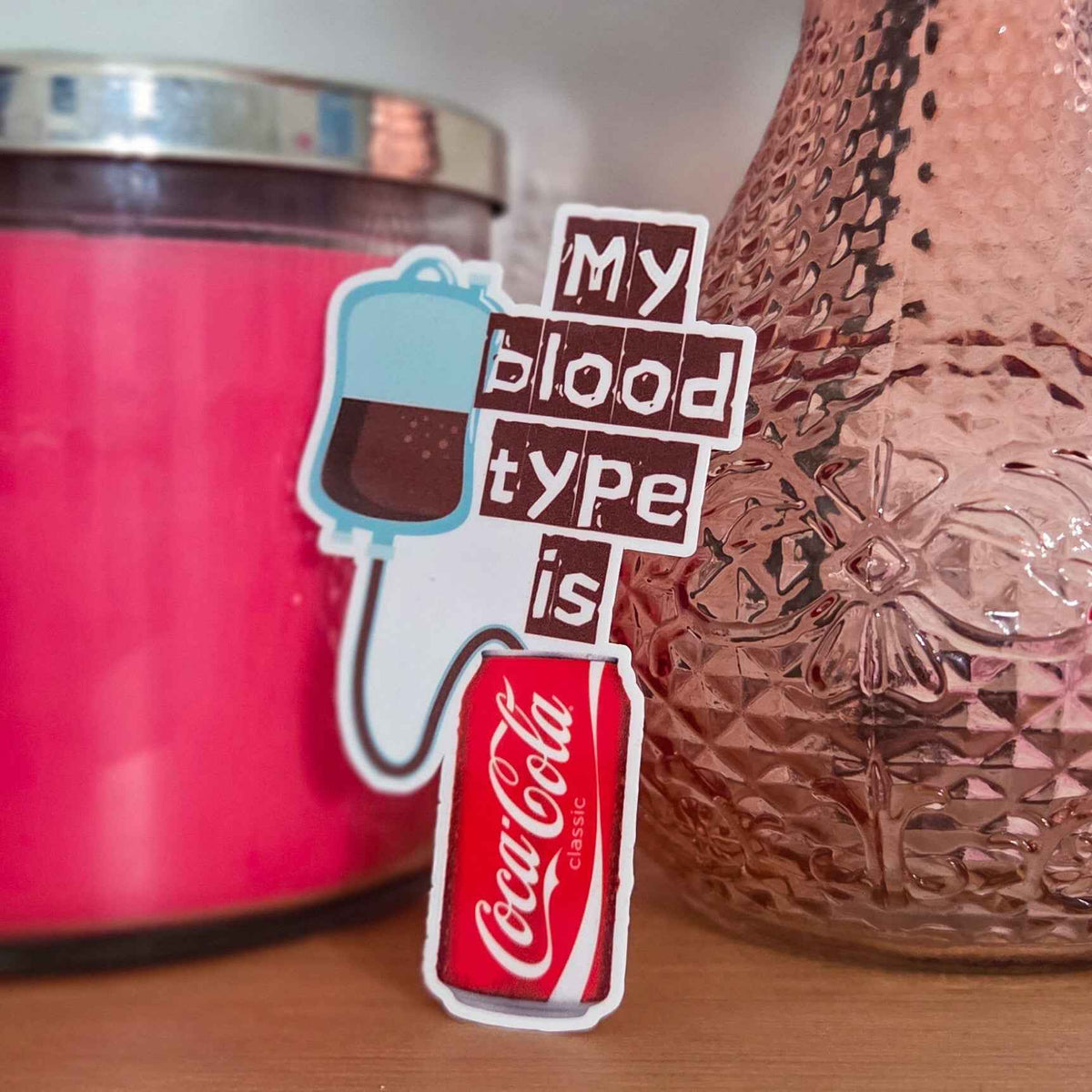 Sticker with 'My blood type is Coke' text next to a pink candle and textured vase.