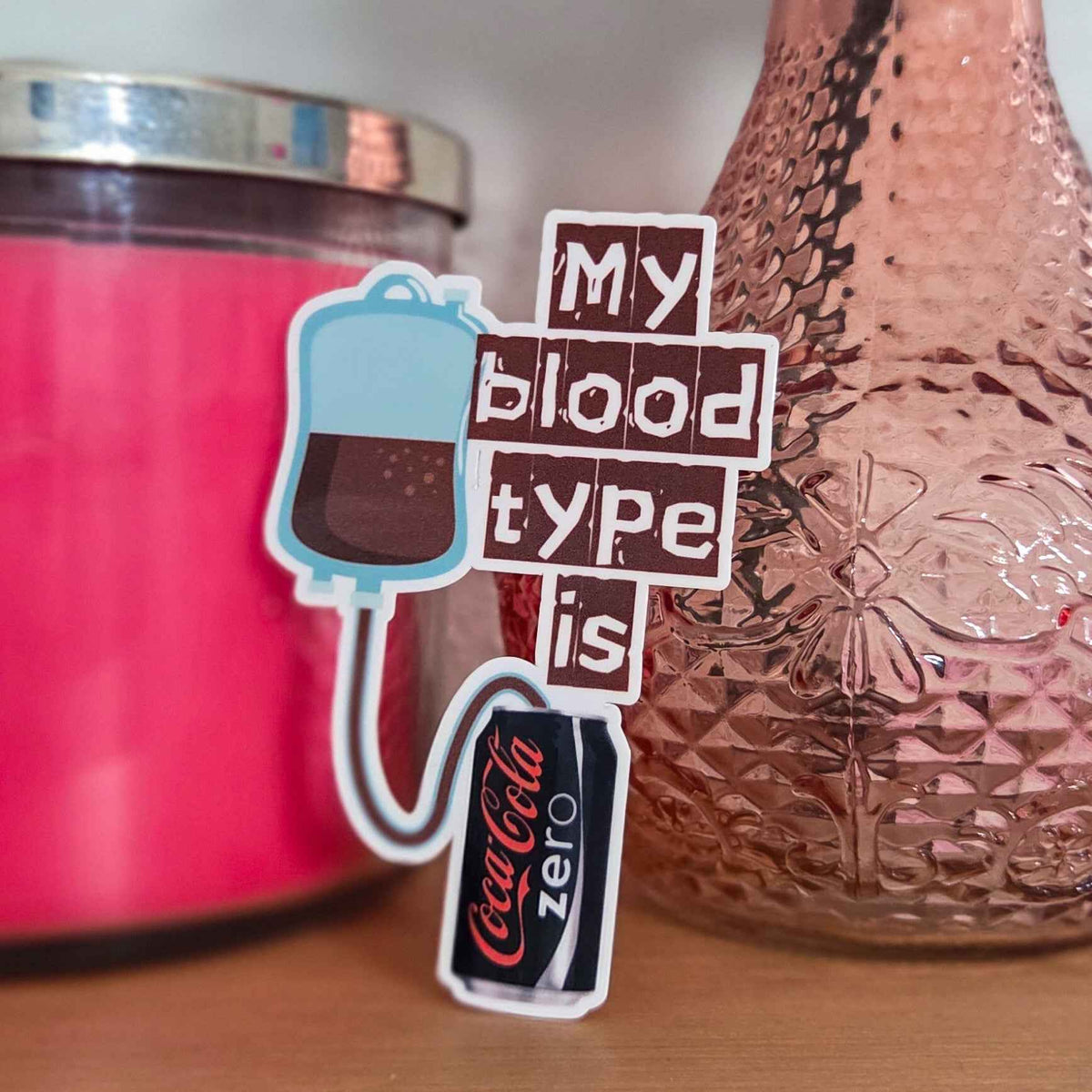 Sticker with 'My blood type is Coke Zero' text next to a pink candle and textured vase.