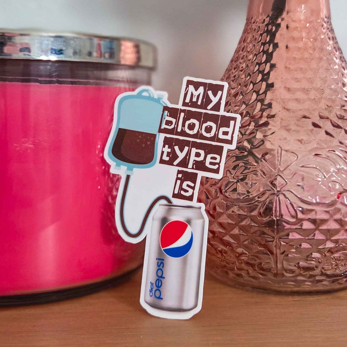 Sticker with 'My blood type is Diet Pepsi' text next to a pink candle and textured vase.