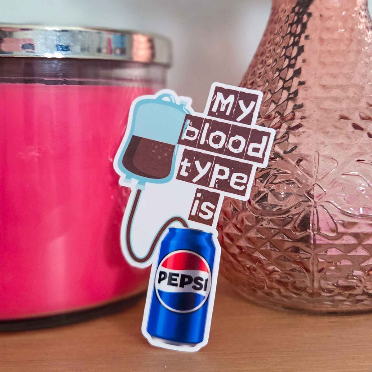 Sticker with 'My blood type is Pepsi' text next to a pink candle and textured vase.