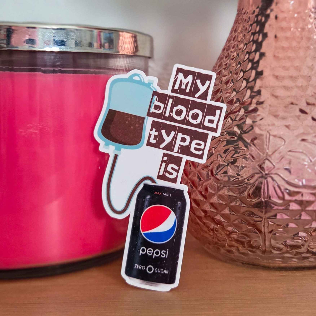 Sticker with 'My blood type is Pepsi Zero' text next to a pink candle and textured vase.
