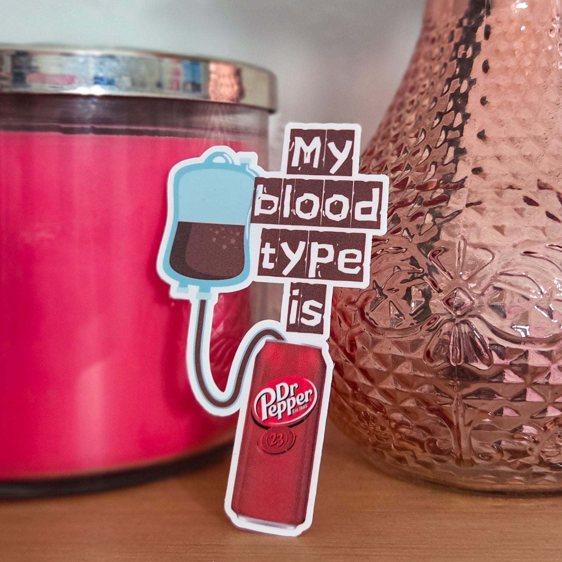 Pink candle, textured vase with flowers, and 'My blood type is' sticker on a wooden surface.