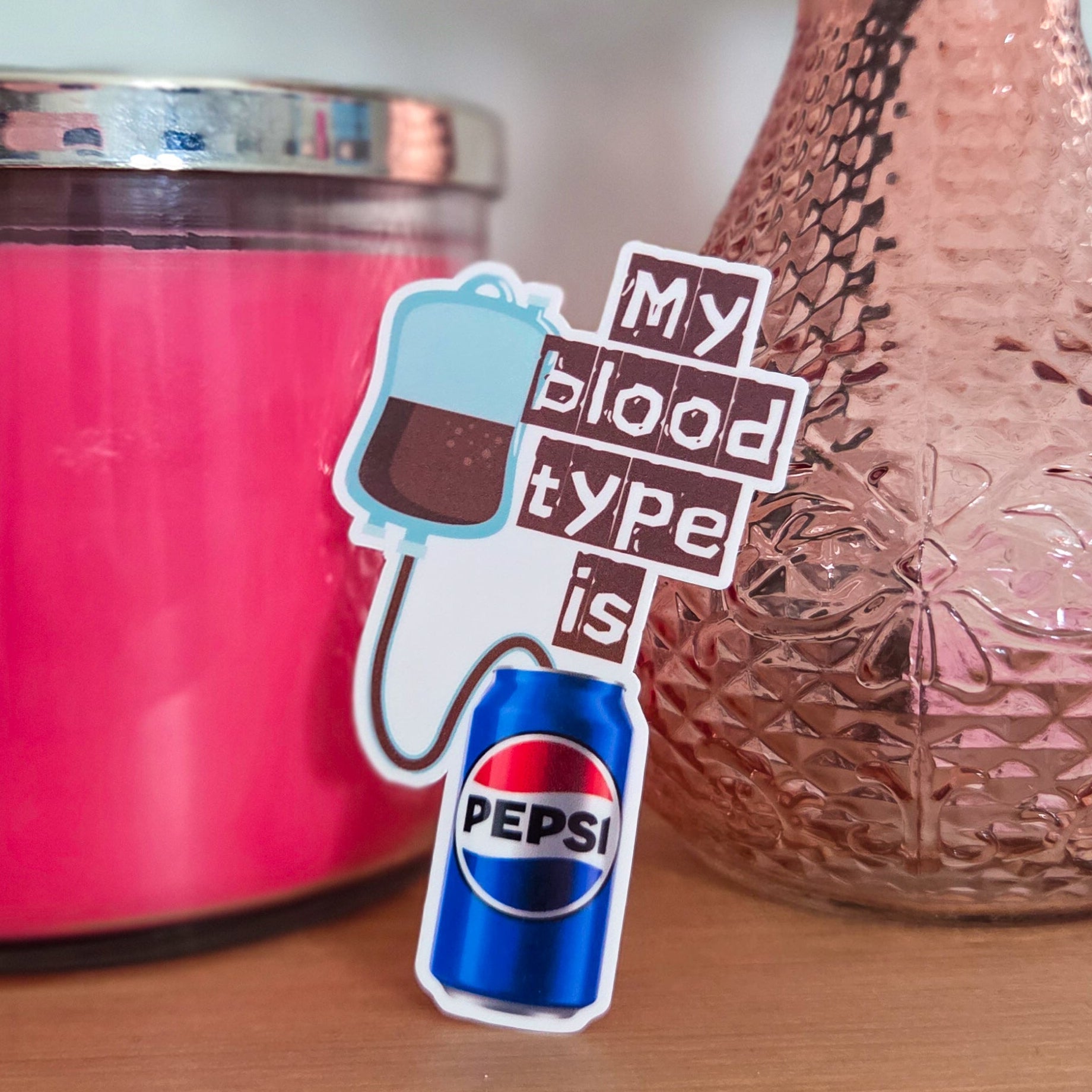 Pink container, vase with flowers, and a sticker with a Pepsi can on a wooden surface.