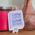 Pink candle, textured vase with peach flowers, and sign on a wooden surface.