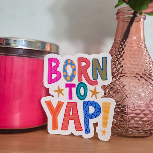 Colorful 'Born to Yap!' sticker on a wooden surface with a pink container and vase in the background.