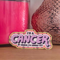 A die-cut sticker with the text 'I'M A CANCER OF COURSE I'M GONNA CRY' in purple lettering, styled with retro pink, yellow, and white borders, resting on a wooden surface in front of pink and metallic decorative objects.