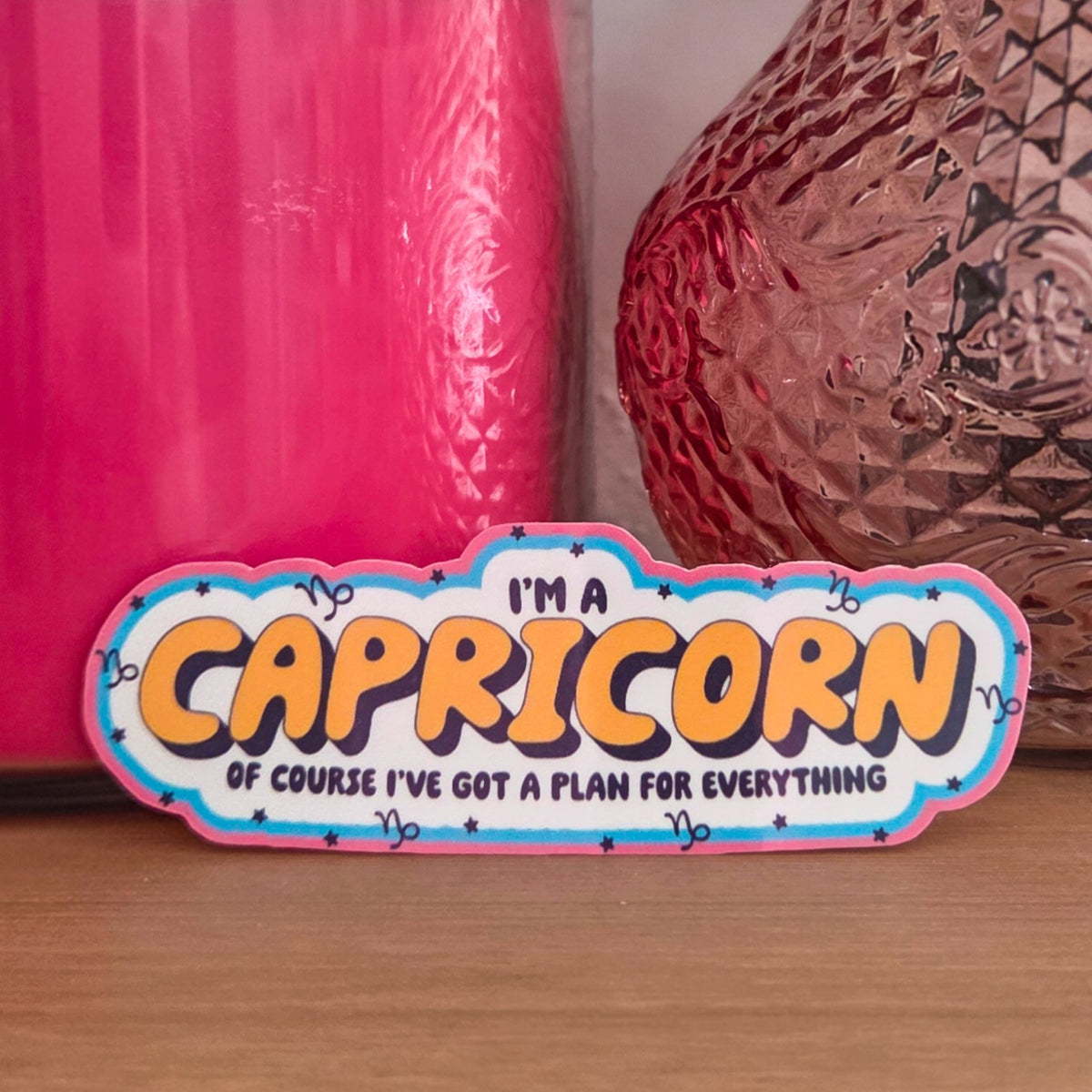 A die-cut sticker with a colorful astrological design that reads 'I'M A CAPRICORN OF COURSE I'VE GOT A PLAN FOR EVERYTHING' resting on a wooden surface.