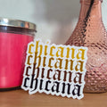 Sticker with 'chicana' text on a wooden surface with a pink vase and candle in the background.