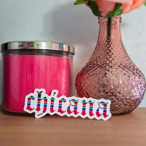 Pink candle, pink vase with flowers, and 'chicana' sticker on a wooden surface.