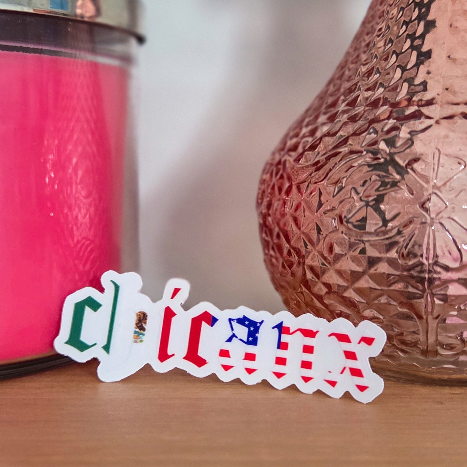 Colorful sticker with 'chicanx' text on a wooden surface next to a pink candle and textured vase.