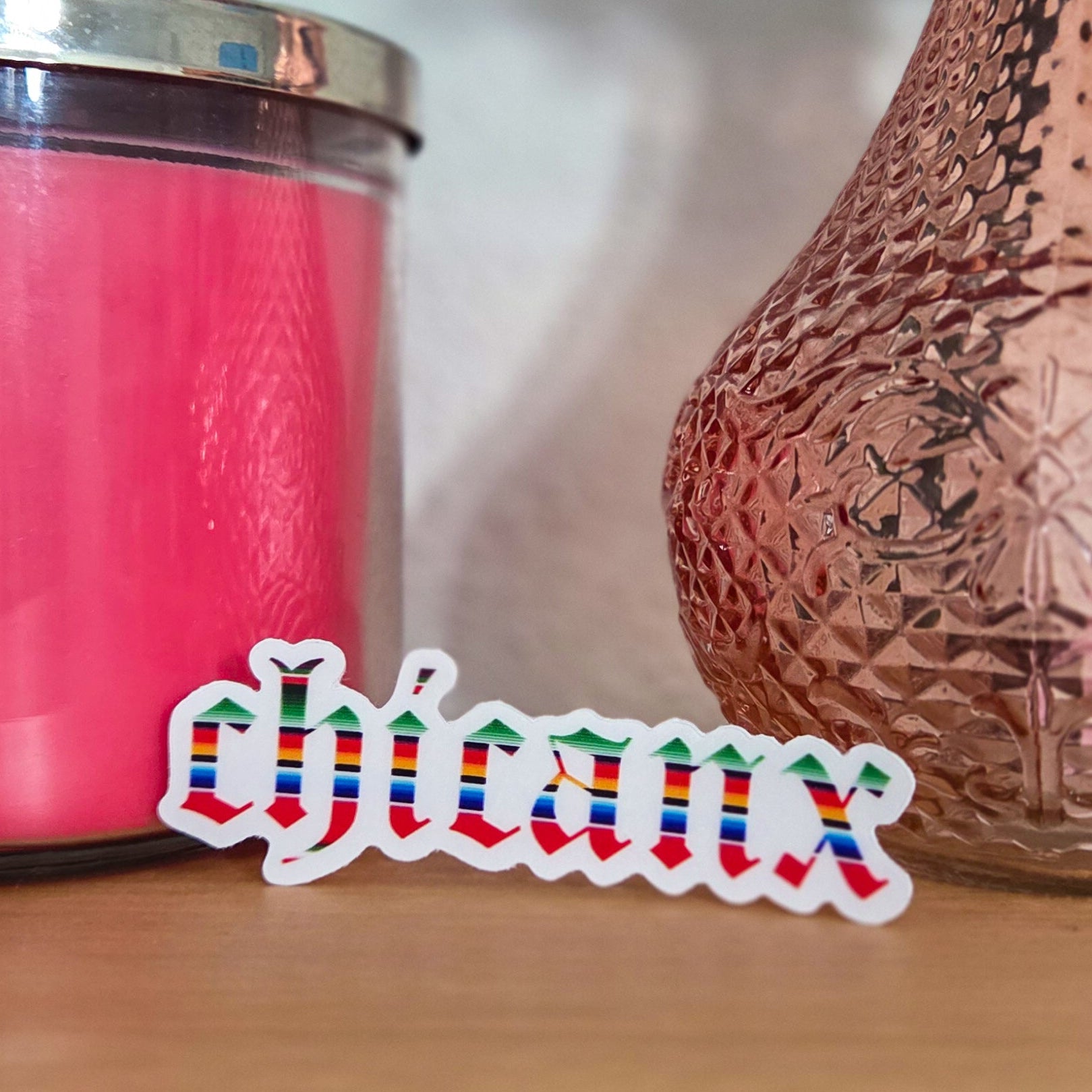 Colorful sticker with 'Chicanx' text next to a pink candle and decorative item on a wooden surface.