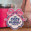 Pink candle, pink vase with a rose, and 'The Cozy Readers Society' sticker on a wooden surface.