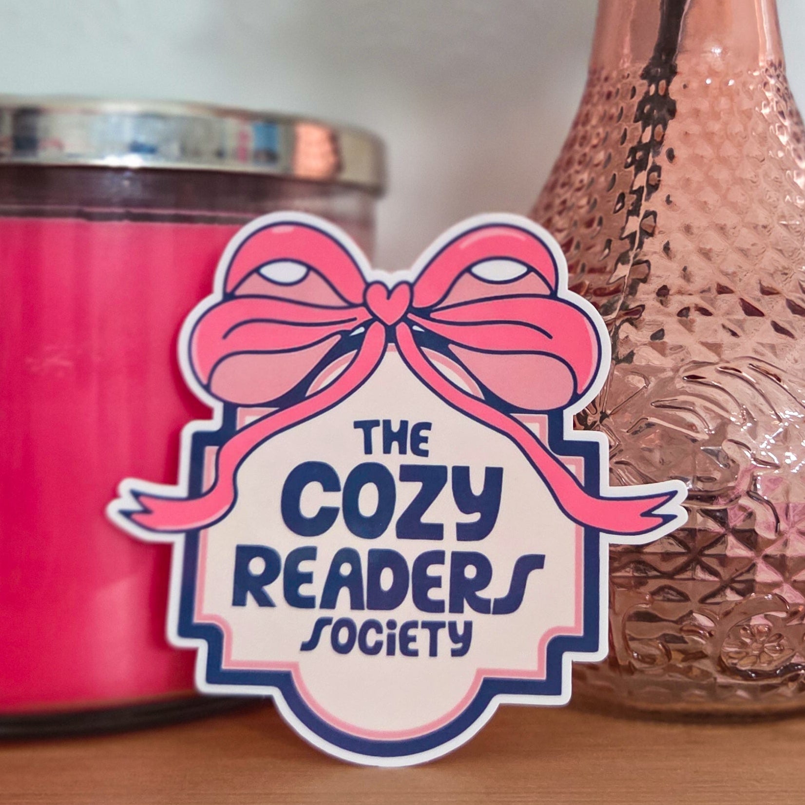 Pink candle, pink vase with a rose, and 'The Cozy Readers Society' sticker on a wooden surface.