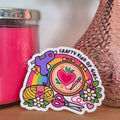 Colorful sticker with text and graphics on a wooden surface next to a pink candle and vase with flowers.