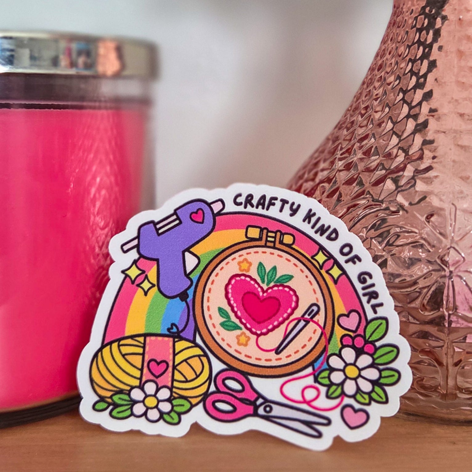 Colorful sticker with text and graphics on a wooden surface next to a pink candle and vase with flowers.
