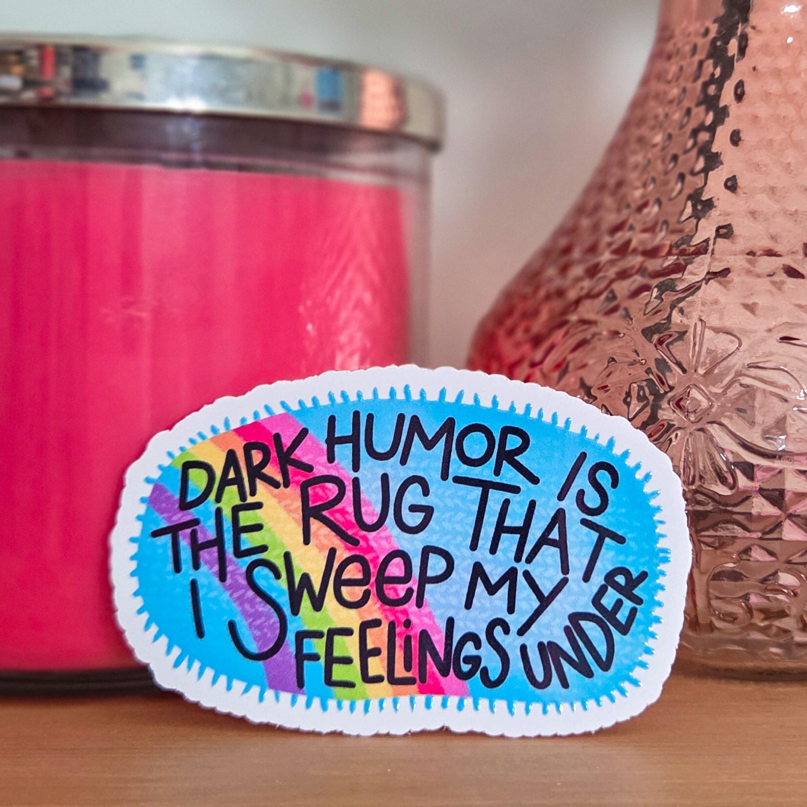Colorful patch with text on a wooden surface with a pink candle and decorative vase in the background.
