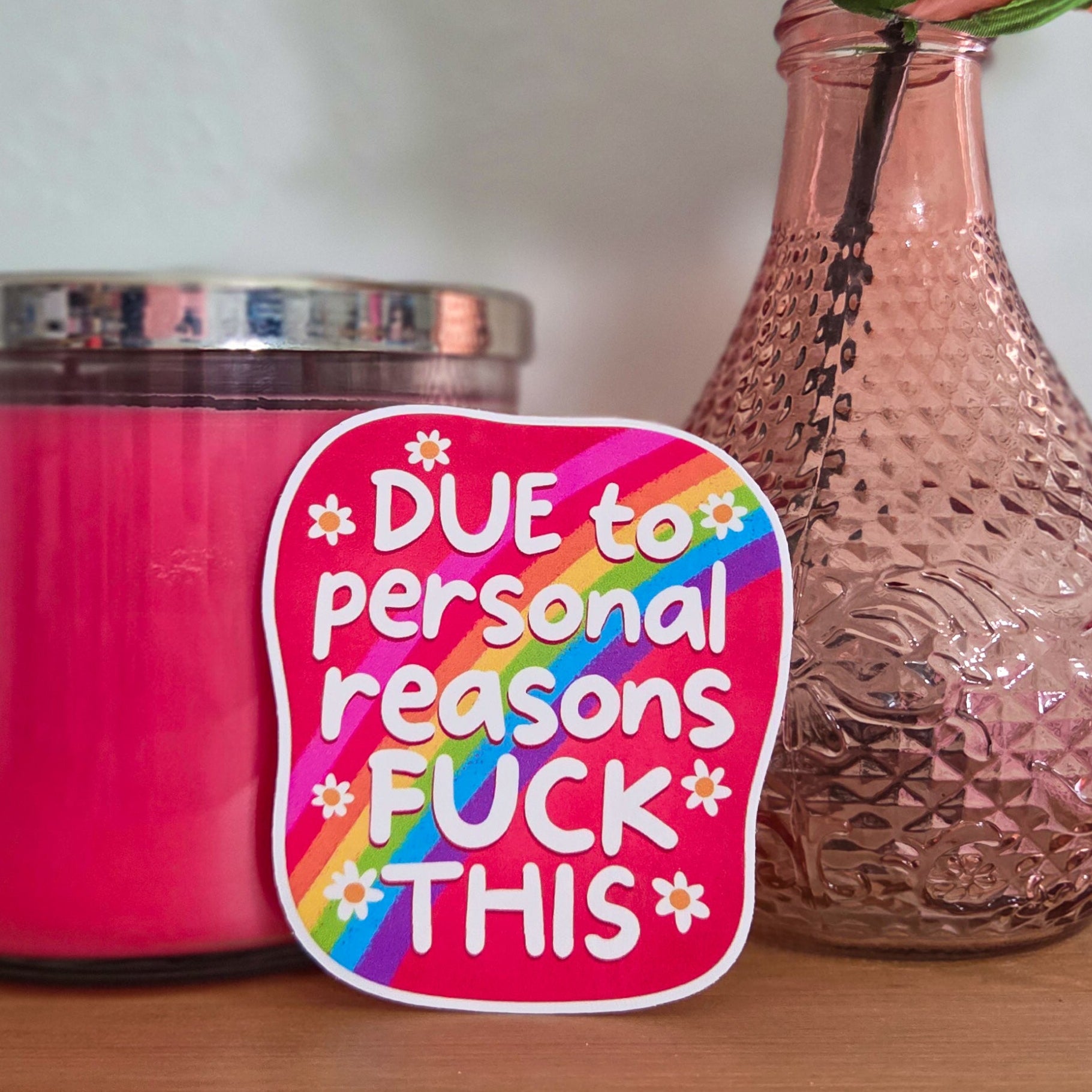 Pink candle, vase with a rose, and colorful sticker on a wooden surface.