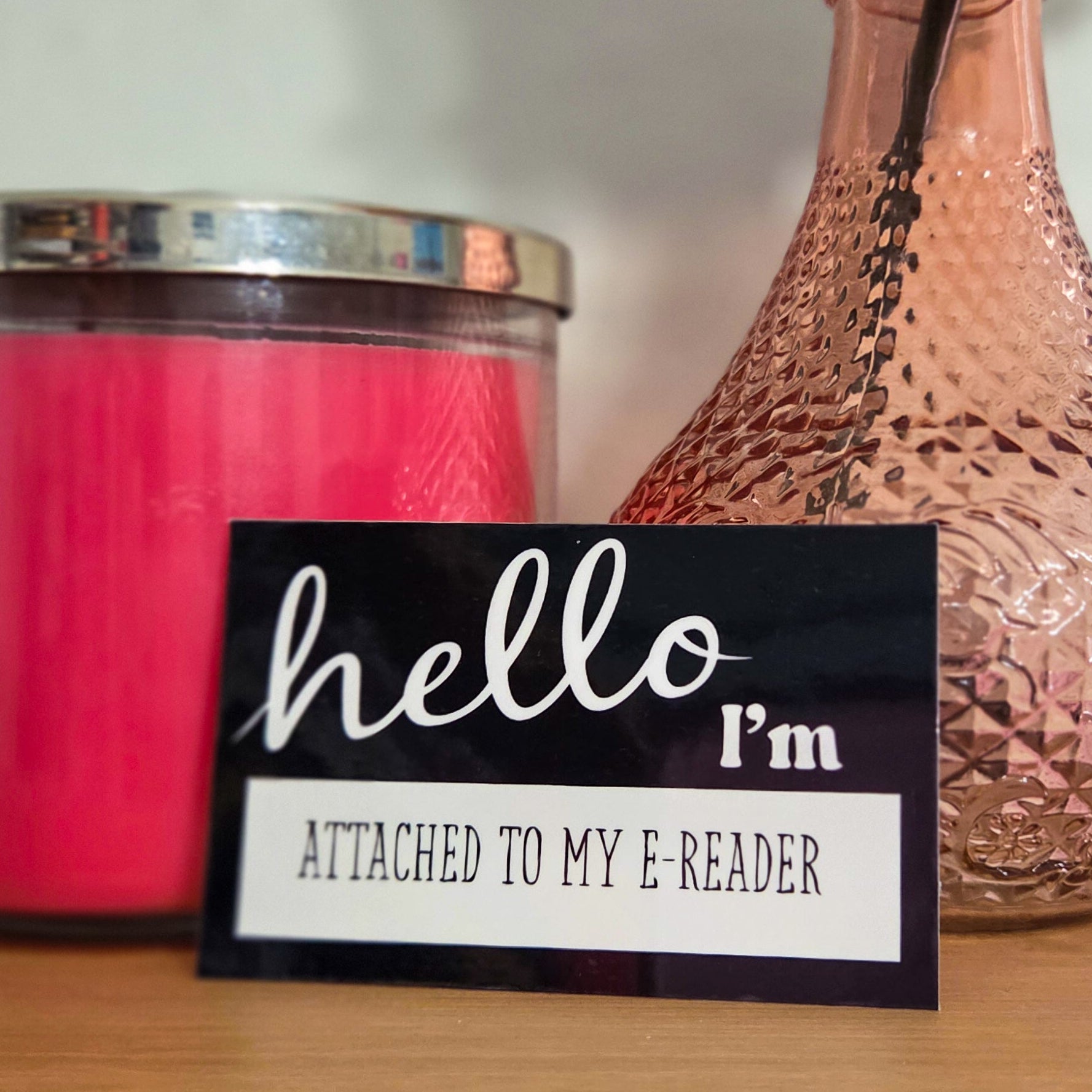 Candle, vase with a rose, and 'hello I'm attached to my e-reader' sticker on a wooden surface.