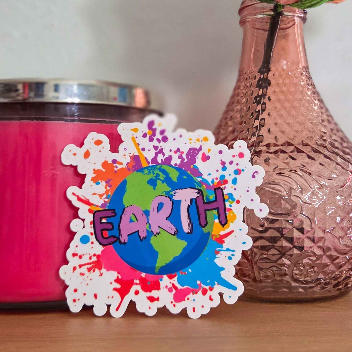Colorful 'EARTH' sticker with a pink candle and textured vase in the background.