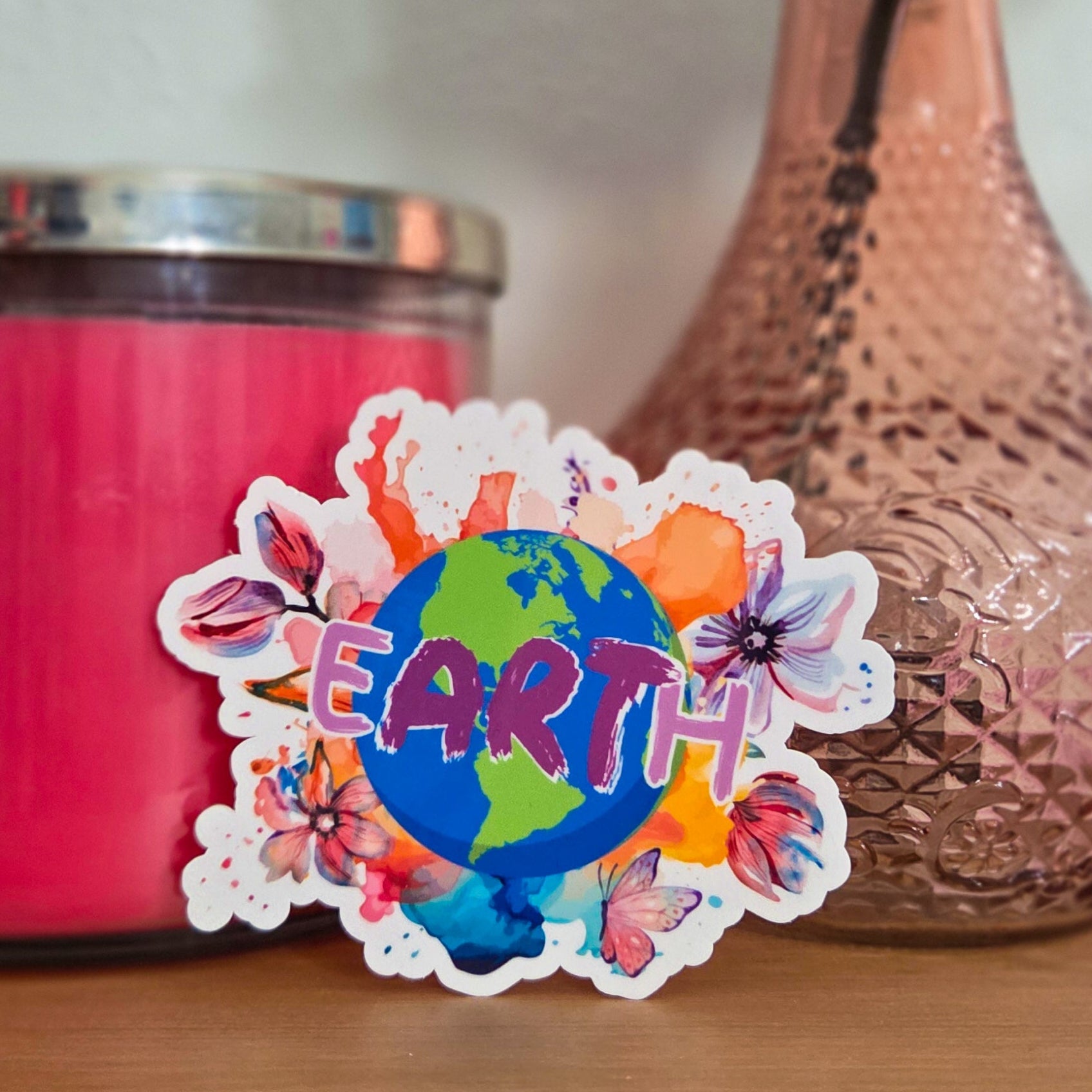 Sticker with 'Earth' design next to a pink candle and vase with flowers on a wooden surface.