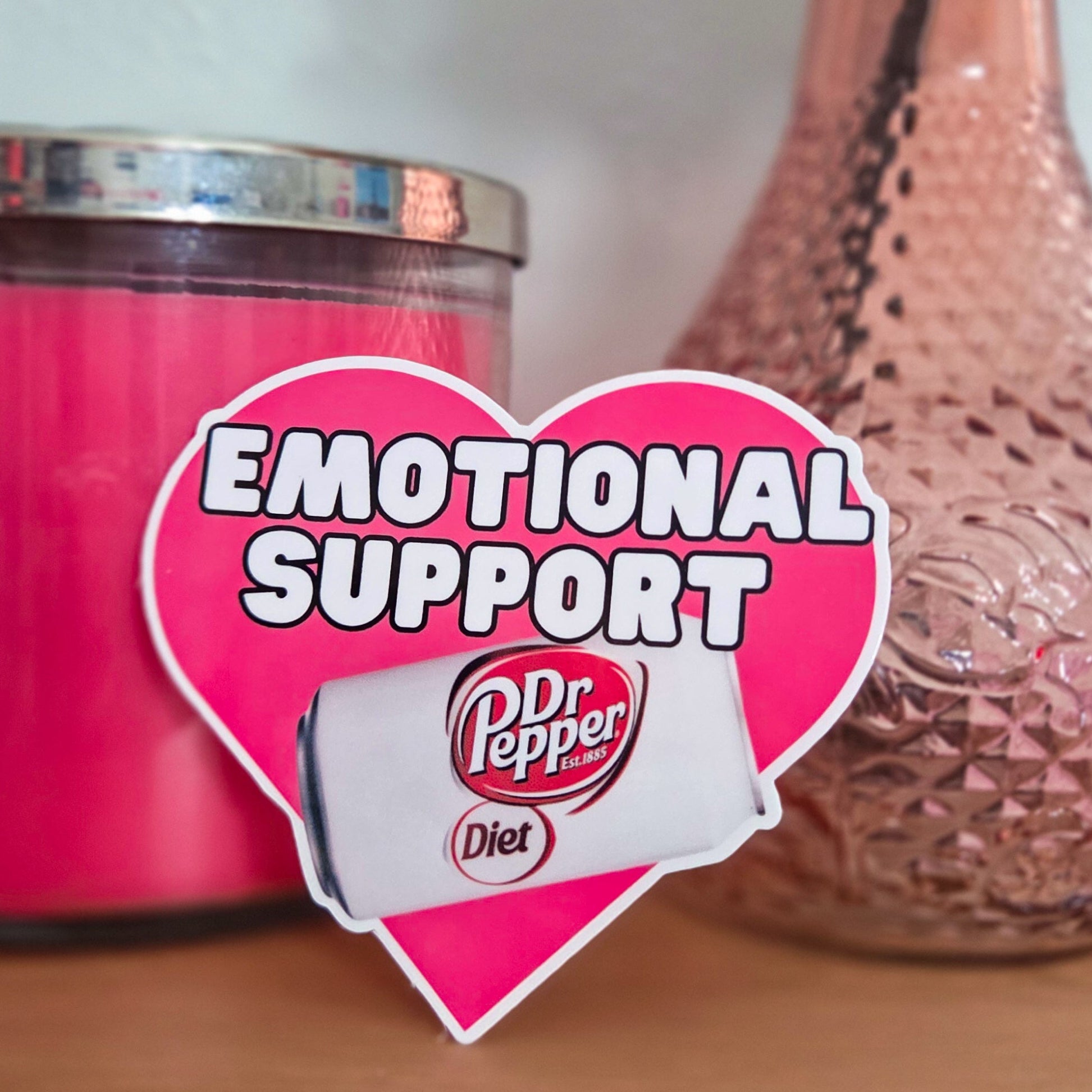 Pink candle labeled 'Emotional Support' with a heart-shaped Diet Dr Pepper label next to a pink vase on a wooden surface.