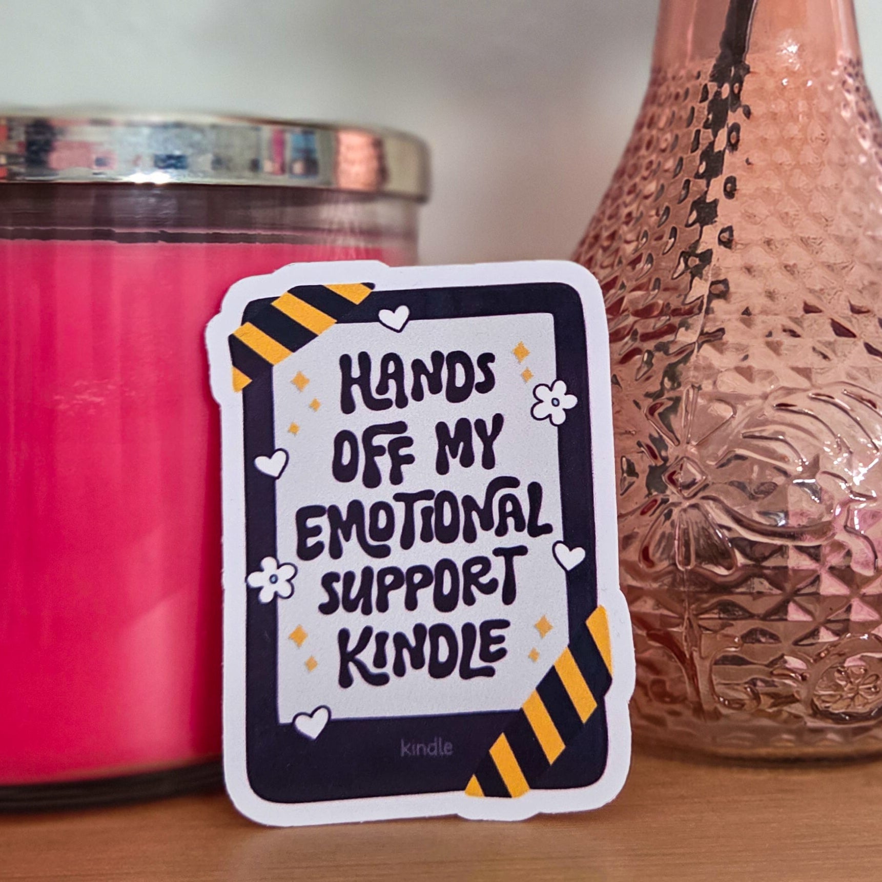 Decorative sticker with text next to a pink candle and vase with flowers on a wooden surface.