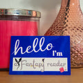 Vase with peach-colored rose, pink candle, and 'hello I'm a fantasy reader' card on a wooden surface.