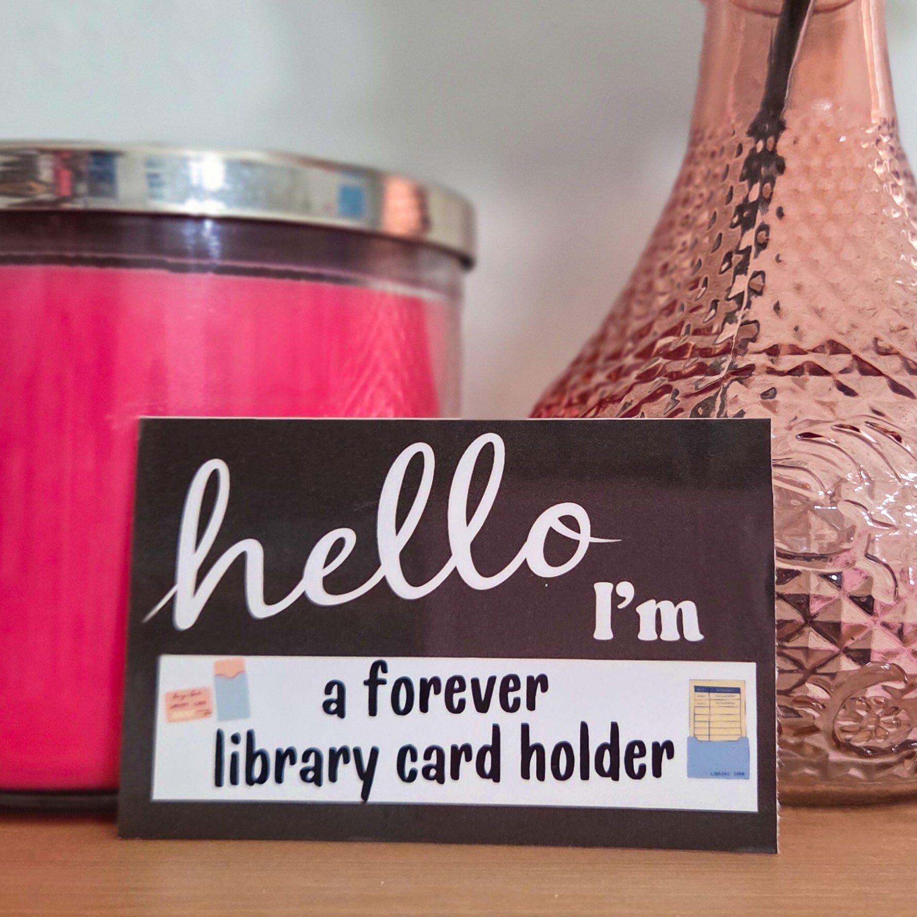 Pink candle, pink vase with a flower, and a sign reading 'hello I'm a forever library card holder' on a wooden surface.