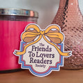 Pink candle, pink vase with a flower, and 'Friends To Lovers Readers' sticker on a wooden surface.