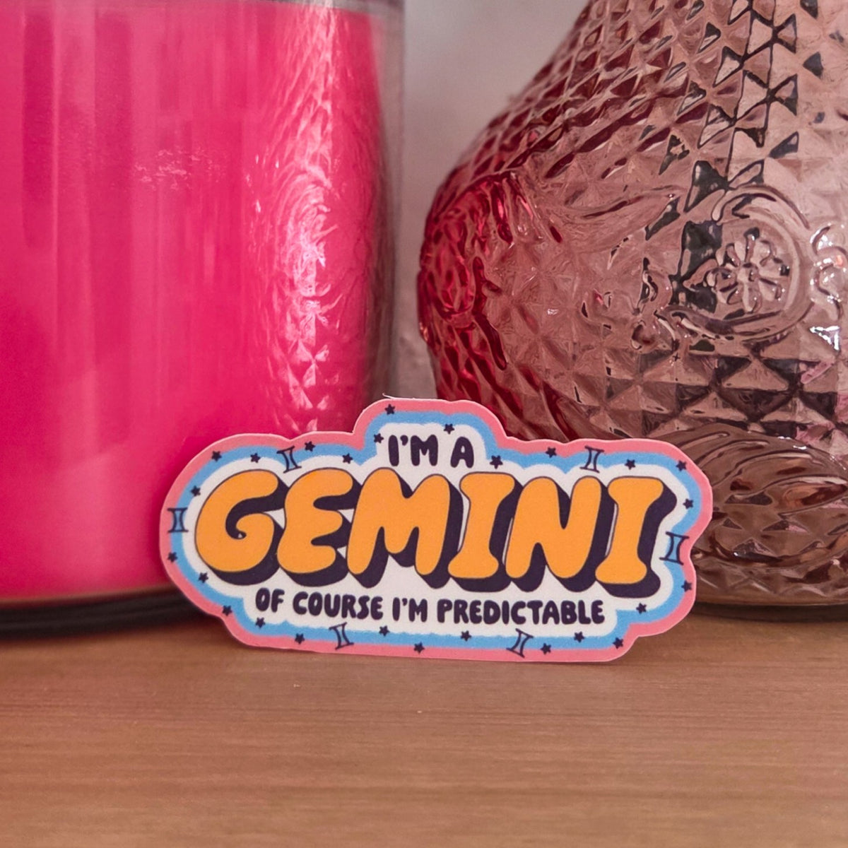 A novelty sticker with the text 'I'M A GEMINI OF COURSE I'M PREDICTABLE' in yellow and black lettering on a pink, blue, and white comic-style background, resting on a wooden surface with a pink candle and a textured metallic vase behind it.
