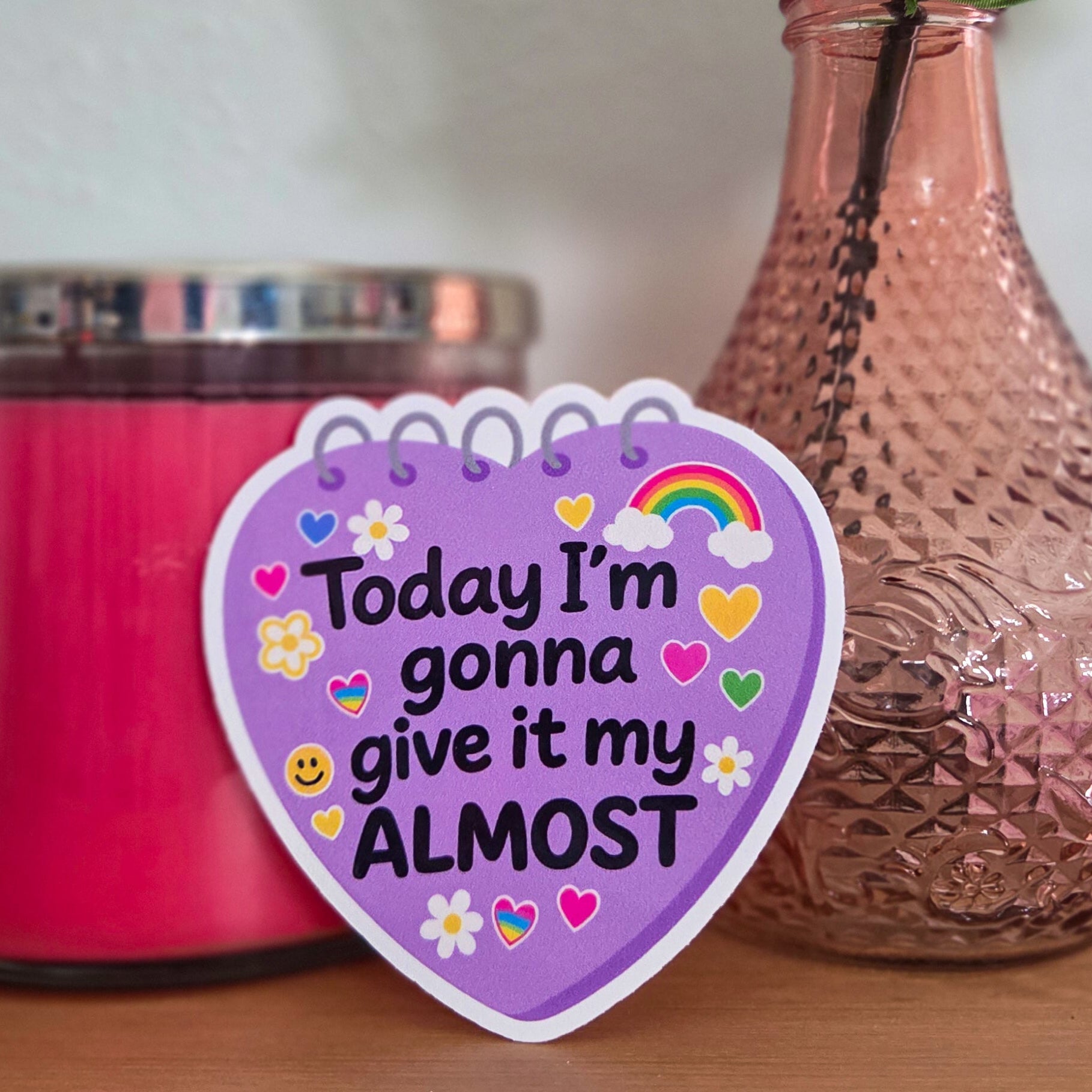 Pink candle, vase with peach roses, and motivational sticker on a wooden surface.