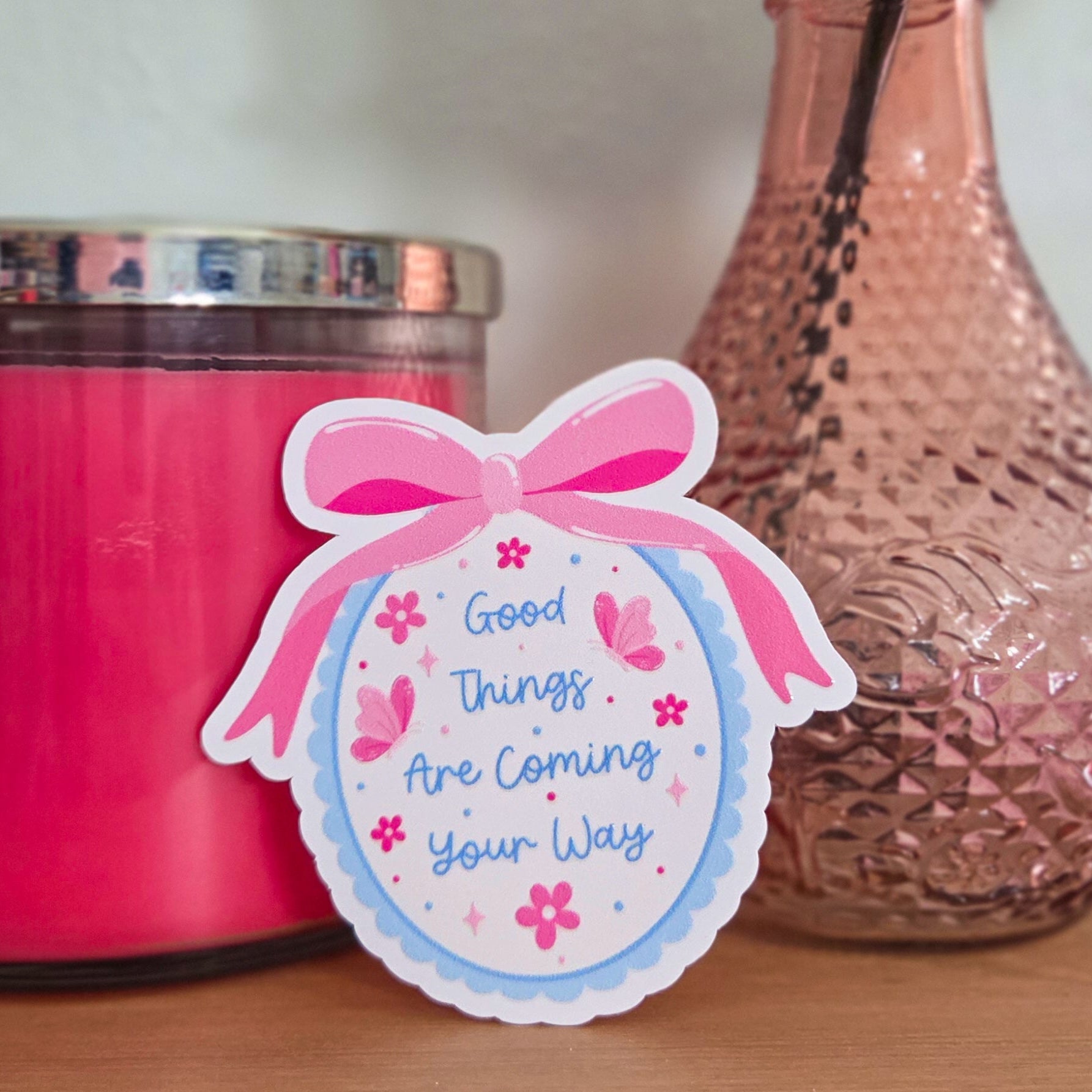 Pink candle, vase with flowers, and an inspirational sticker on a wooden surface.