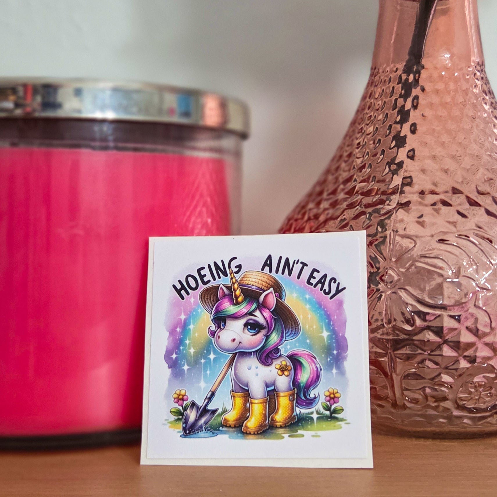 Pink candle, pink vase with a flower, and a card with a colorful unicorn design on a wooden surface.