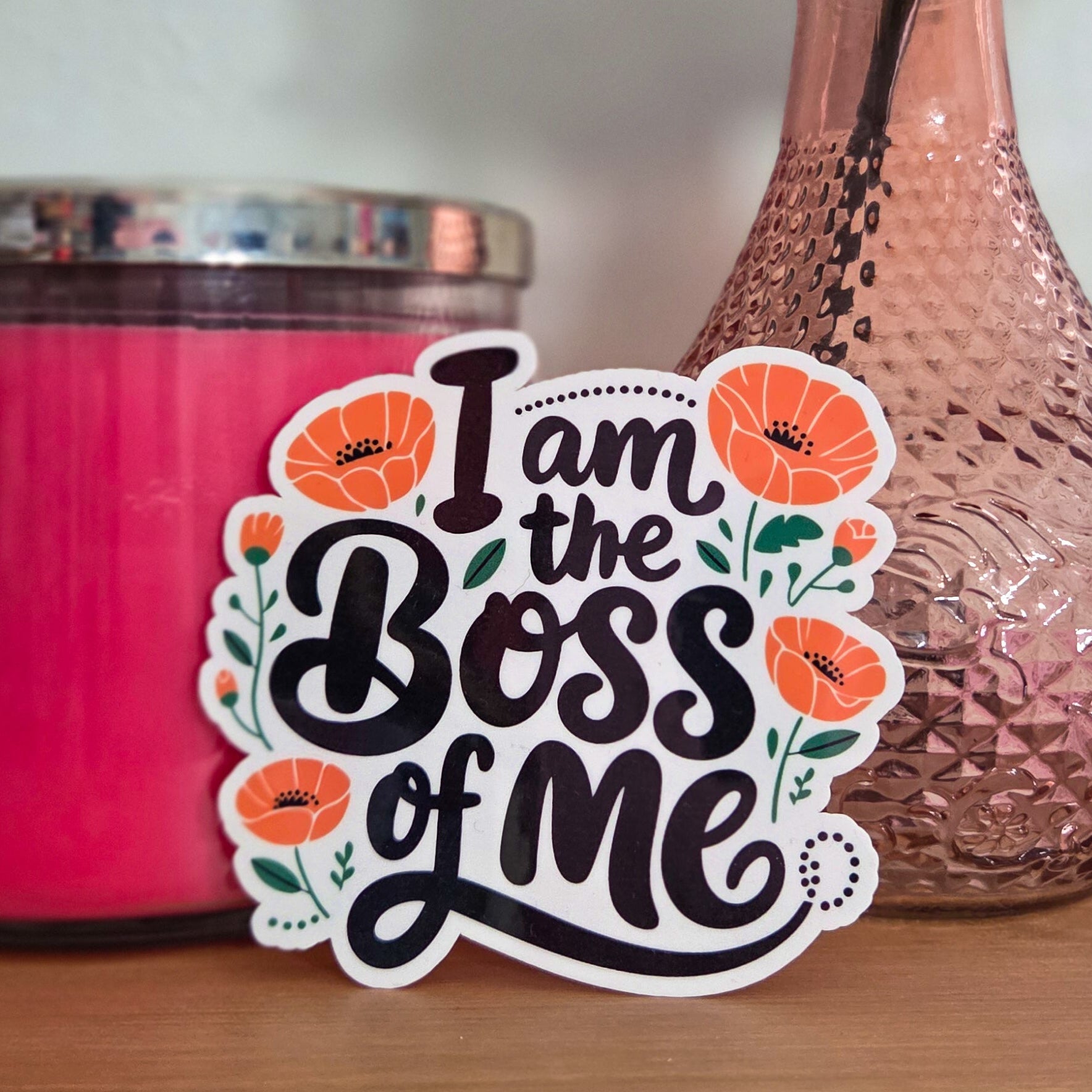 Sticker with 'I am the boss of me' text on a pink candle and vase with flowers.