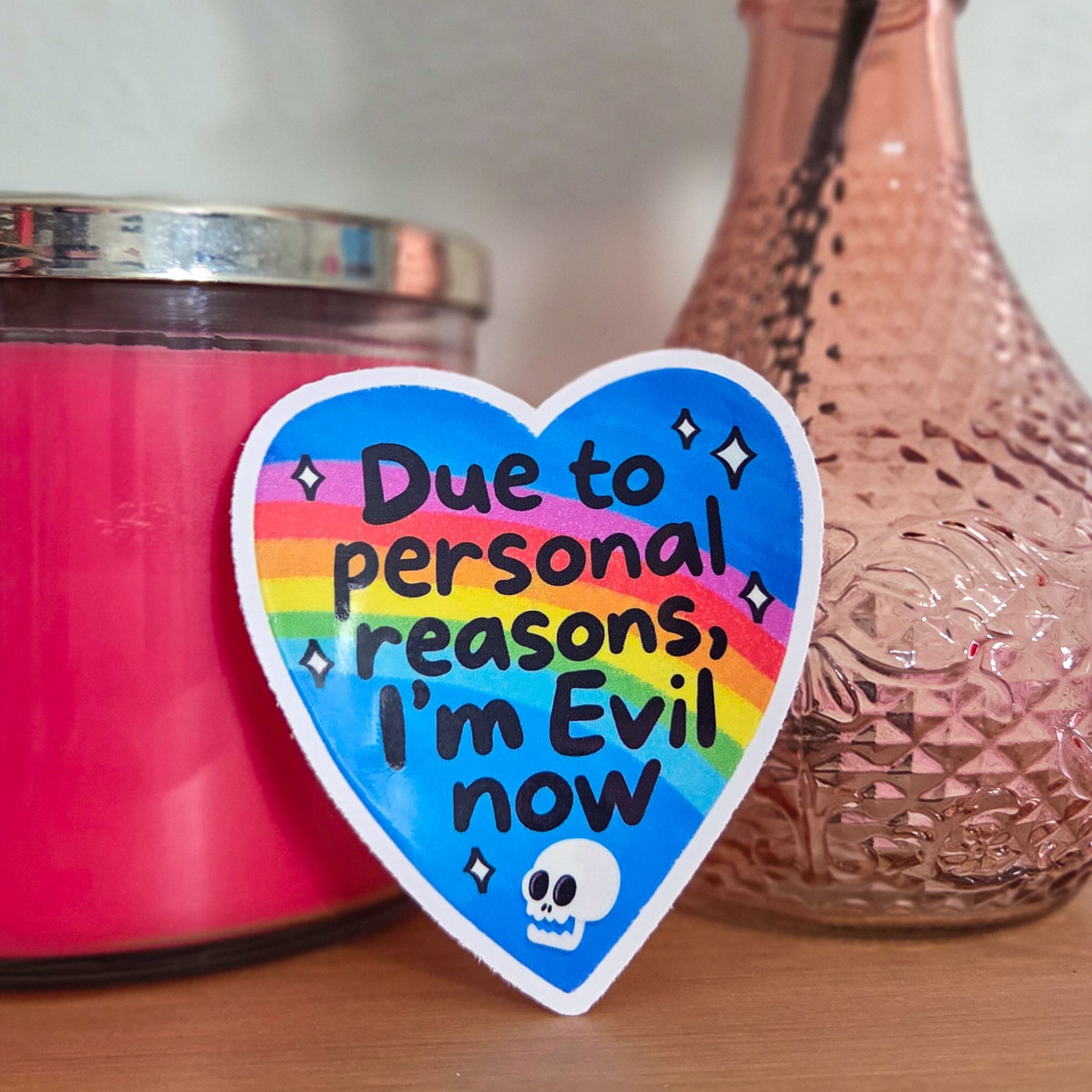 Heart-shaped sticker with text next to a pink candle and vase on a wooden surface.
