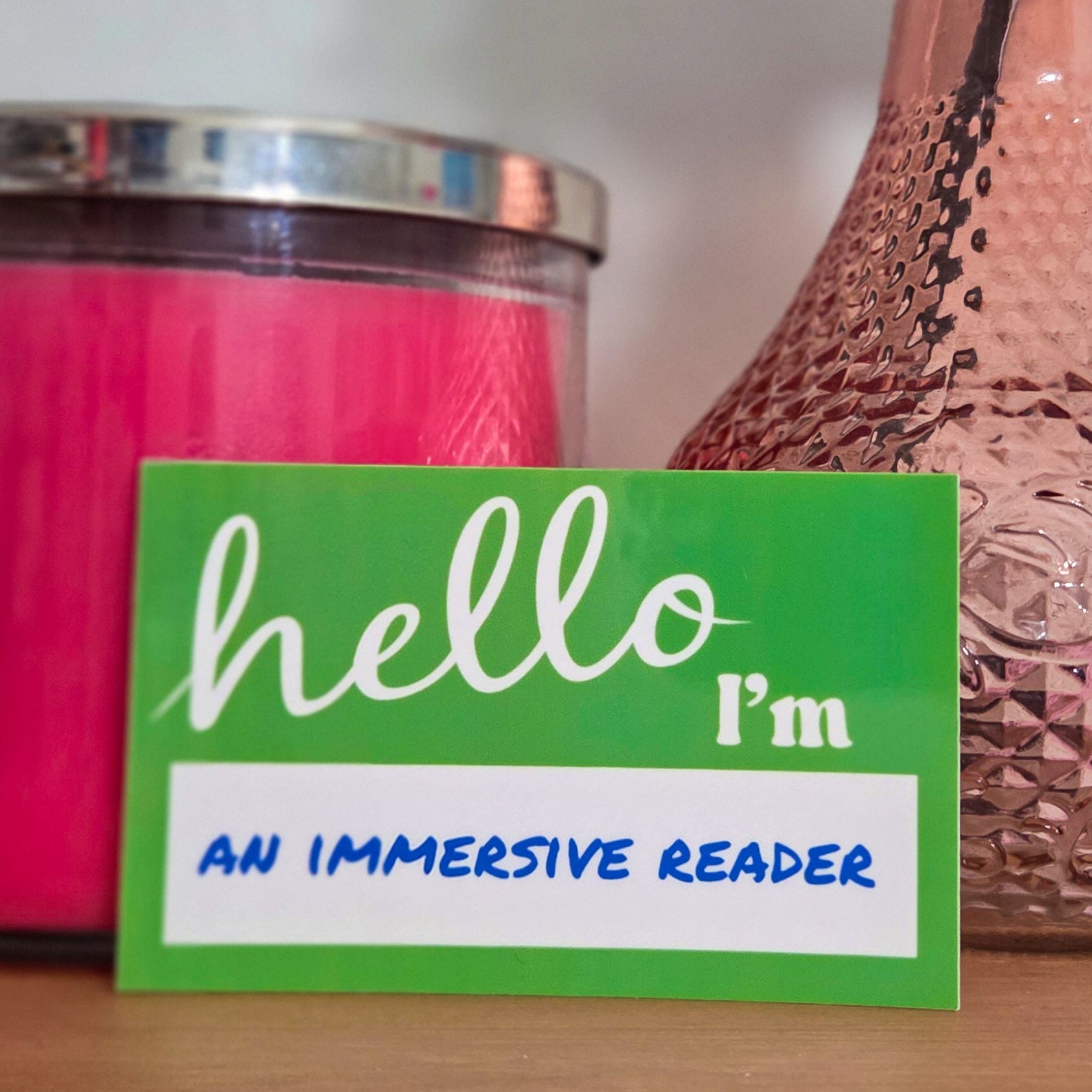 Green sticker with 'hello, I'm an immersive reader' text on a wooden surface with a pink vase and candle in the background.