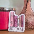 Sticker with 'Just Let Me Read' text on a wooden surface with a pink candle and vase in the background.