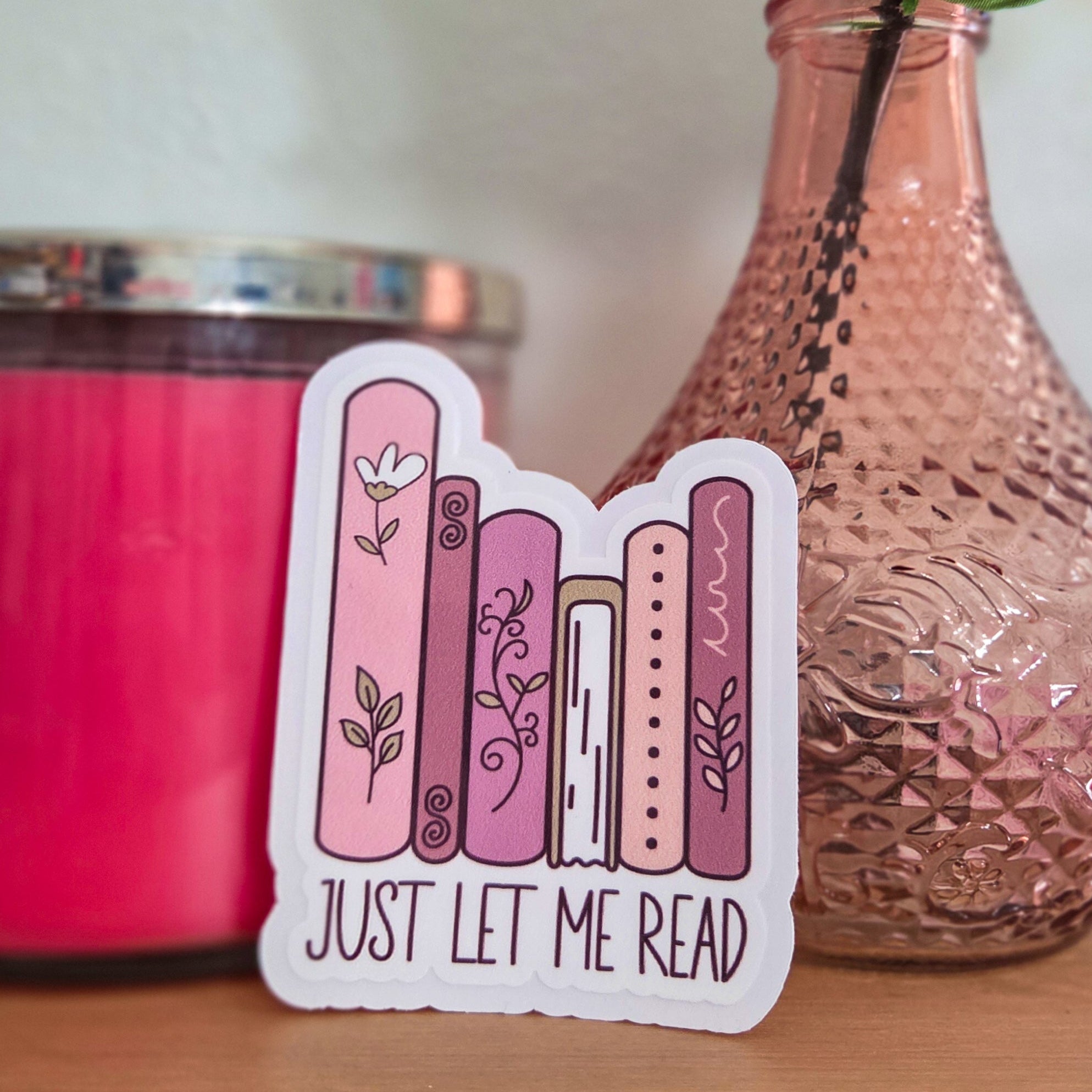Sticker with 'Just Let Me Read' text on a wooden surface with a pink candle and vase in the background.