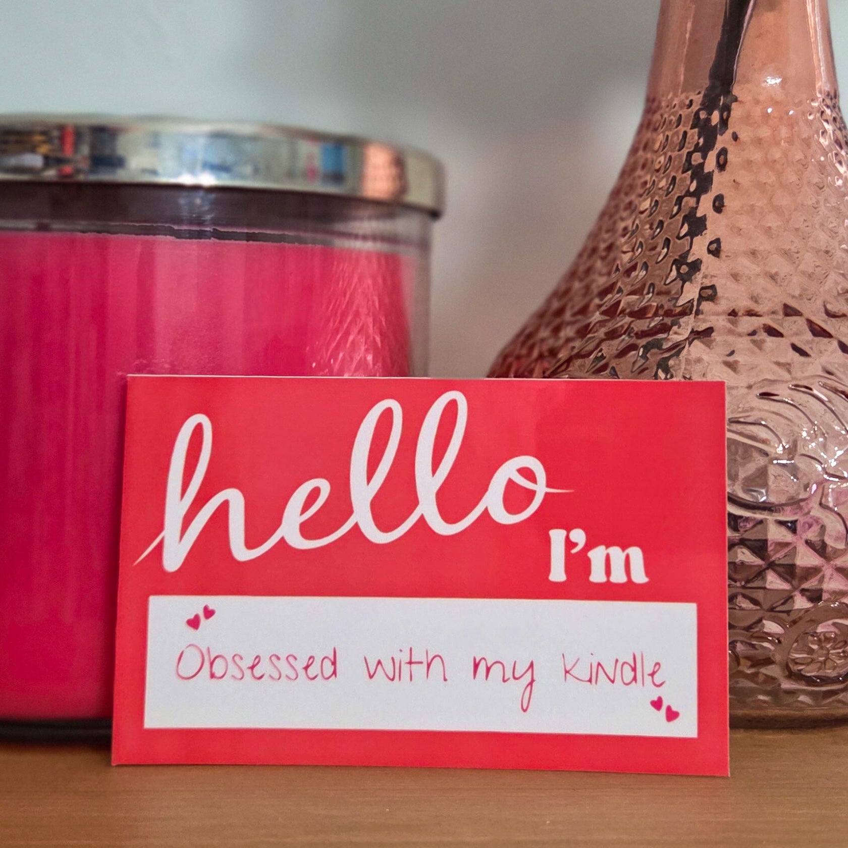 Pink candle, textured vase with a rose, and 'hello I'm obsessed with my kindle' card on a wooden surface.