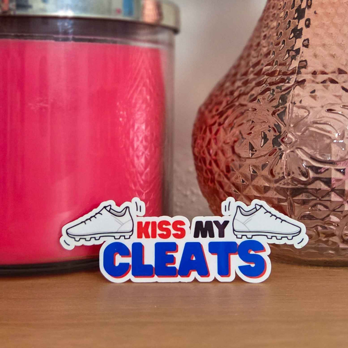 Pink candle and decorative vase with 'Kiss My Cleats' sticker on a wooden surface