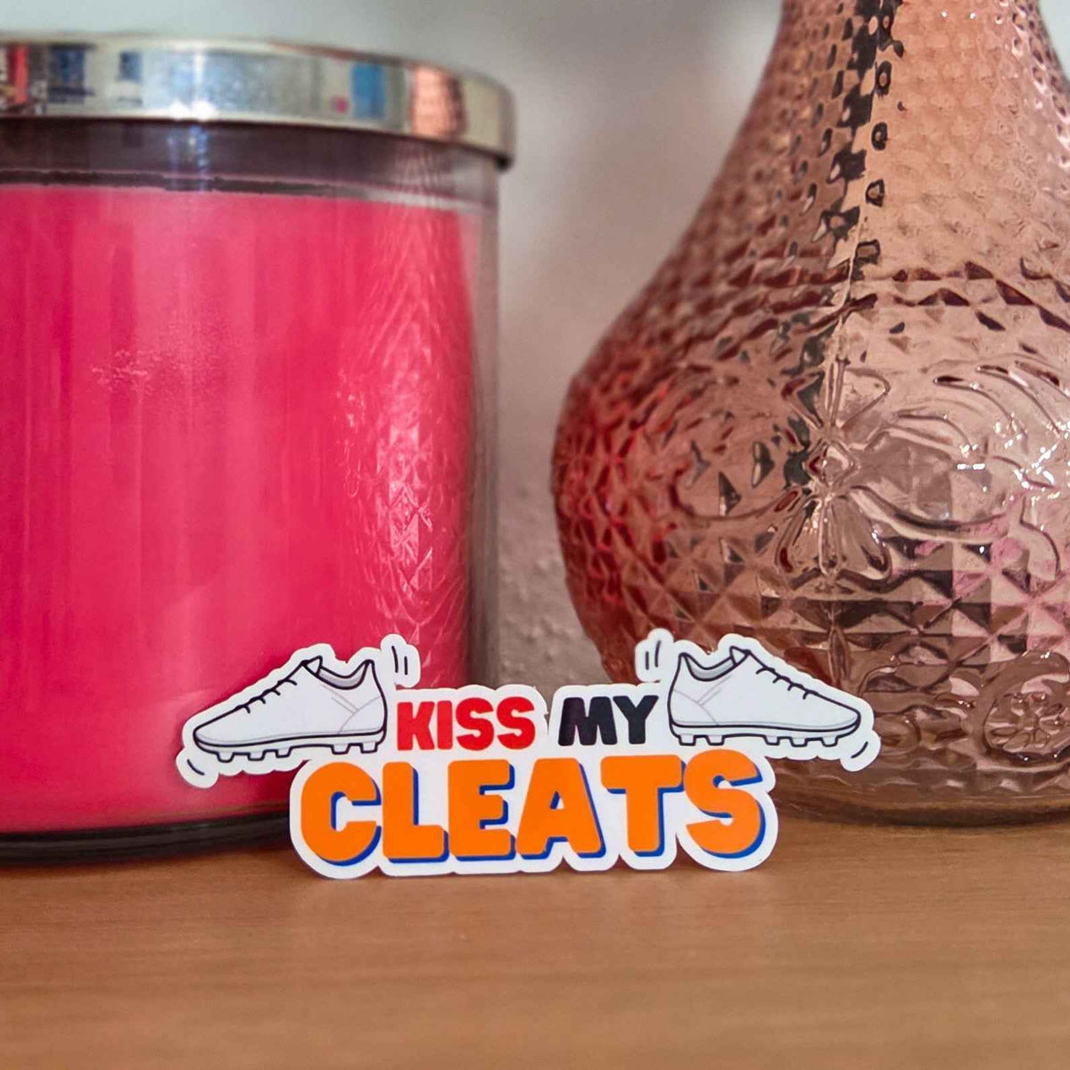 Pink candle and decorative vase with 'Kiss My Cleats' sticker on a wooden surface